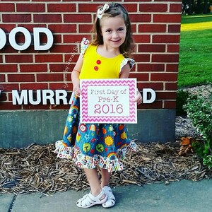 May include: A young girl in a yellow shirt and a colorful skirt holds a pink sign that says "First Day of Pre-K 2016".