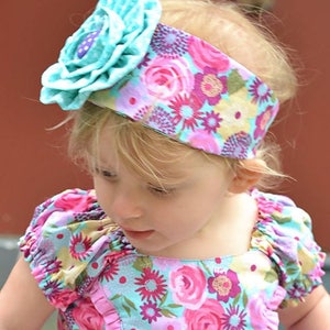 Op de afbeelding: Een jong meisje draagt ​​een hoofdband met een grote turquoise bloem en een bloemenprint. De hoofdband is gemaakt van stof en heeft een roze en blauwe bloemenprint. Het meisje draagt ​​een jurk met een vergelijkbare bloemenprint.