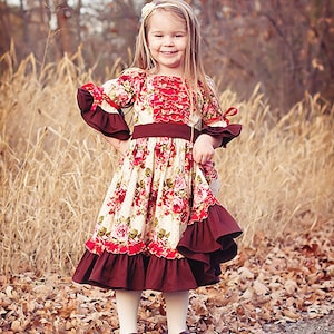 Op de afbeelding: Een jong meisje draagt een jurk met bloemenprint met een bruine ruche en bruine schoenen. Ze staat in een veld met hoog gras en gevallen bladeren.