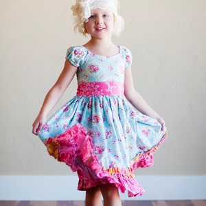 May include: A young girl wearing a light blue and pink floral dress with a white headband. The dress has a pink sash and a ruffled hem.