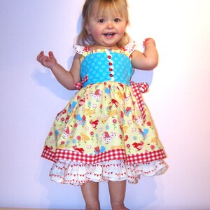 Puede incluir: Una niña pequeña con un vestido azul, amarillo y rojo con un borde de volantes blancos. El vestido tiene un estampado floral con pájaros y un borde de cuadros rojos y blancos. La niña sonríe y lleva un lazo rojo en el pelo.
