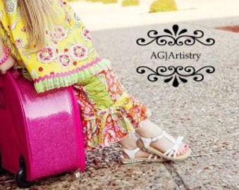 May include: A young girl sits on a pink heart-shaped suitcase with wheels. She is wearing a yellow and green floral dress with a pink bracelet and white sandals.