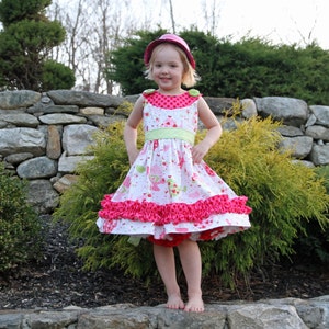 Puede incluir: Una niña pequeña con un vestido rosa y blanco con una cinta verde y un sombrero rosa. El vestido tiene un dobladillo con volantes y un patrón de lunares blancos. La niña está de pie frente a una pared de piedra con plantas verdes.