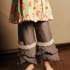 Op de afbeelding: Een jong meisje draagt een groene jurk met bloemenprint met een geruite zoom en grijze spijkerbroek met kantversiering en meerdere ruches. De broek heeft een klein wit label met de tekst "Rooster Threads" op het linkerbeen.