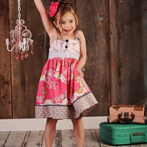 May include: A young girl wearing a pink and white dress with a carousel print and a black bow. She is wearing yellow shoes and has a pink flower in her hair. The dress has a white lace trim and a polka dot bottom.
