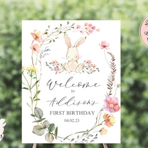 May include: A printable welcome sign for a first birthday party. The sign features a watercolor illustration of a white bunny rabbit surrounded by pink and yellow flowers. The text on the sign reads "Welcome to Addison's First Birthday 04.02.23".