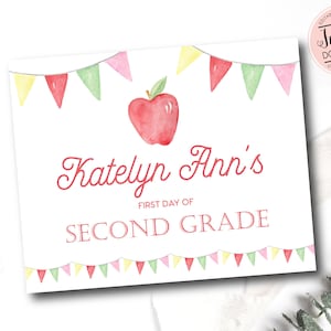 以下が含まれることがあります： 赤いリンゴとカラフルな旗飾りで飾られた、印刷可能なサイン。「Katelyn Ann's First Day of Second Grade」と書いてあります。