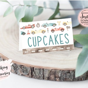 May include: A white card with the word "CUPCAKES" in teal letters, featuring watercolor illustrations of race cars. The card includes a road design at the bottom and is set on a wooden surface.
