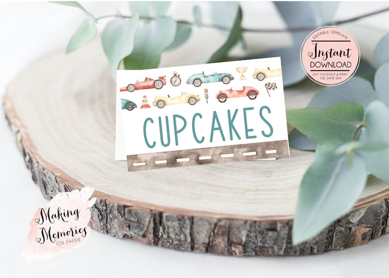 May include: A white card with the word "CUPCAKES" in teal letters, featuring watercolor illustrations of race cars. The card includes a road design at the bottom and is set on a wooden surface.