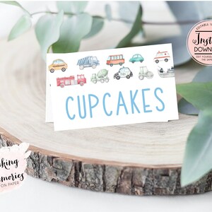 Op de afbeelding: Een witte kaart met het woord "CUPCAKES" in lichtblauw, met aquarel illustraties van diverse voertuigen. De kaart ligt op een houten schijf, met de tekst "Making Memories ON PAPER".