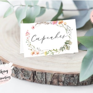 K&ouml;nnte beinhalten: Druckbarer Schild mit einem floralen Kranzdesign und dem Text "Cupcakes". Das Schild befindet sich auf einer Holzscheibe mit gr&uuml;nen Bl&auml;ttern.