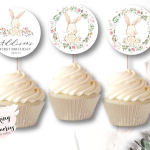May include: Three cupcake toppers featuring a watercolor illustration of a bunny rabbit in a floral wreath. The toppers are printed on white cardstock and have a pink and green color scheme. The text on the topper reads "Addison's First Birthday 04.02.23".