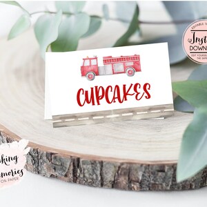 May include: Printable food label with a red fire truck illustration and the word "CUPCAKES" in red text. The label is on a wooden surface with a white background.