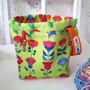 Könnte beinhalten: Ein lindgrüner Stoffbeutel mit Kordelzug und einem bunten Blumenmuster aus roten, blauen und orangefarbenen Blüten. Die Tasche hat einen orangefarbenen Henkel mit einem kleinen weißen Etikett, das eine Reihe kleiner bunter Herzen zeigt, sowie einen roten Clip.