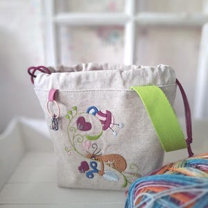 Könnte beinhalten: Ein beigefarbener Leinenbeutel mit Kordelzug, verziert mit einer Stickerei eines braunen Katers, lila Herzen und einer blauen Schere. Die Tasche verfügt über einen hellen lindgrünen Stoffgriff, eine dunkelviolette Kordel und eine kleine rosa Schlaufe mit Metallclips.