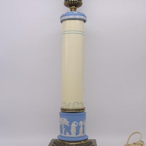 May include: A decorative table lamp with a cream-colored column and blue accents. The lamp features a detailed brass base and top, with a blue base adorned with white relief figures. The lamp is approximately 30 inches tall.