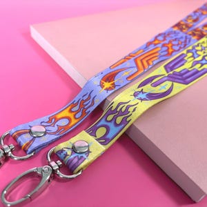 Op de afbeelding: Twee stoffen keycords met levendige grafische prints. De ene band toont oranje en rode vlammen op een lichtpaarse achtergrond, terwijl de andere paarse vlammen op een gele achtergrond heeft. Beide zijn voorzien van zilverkleurige metalen draaibare karabijnhaken.