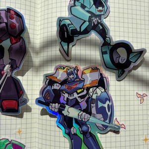 May include: A selection of holographic stickers depicting cartoon robot characters in different poses. The stickers display a variety of colours, including blue, purple, and orange, with a shiny, iridescent finish. The robots are shown with detailed designs and dynamic action poses.