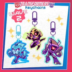 Op de afbeelding: Drie Transformers Wave 2 sleutelhangers. De sleutelhangers tonen stripachtige robotkarakters in blauw, geel en paars. Elke sleutelhanger heeft een metalen sluiting. De afbeelding bevat het Transformers-logo en de tekst "Keychains".