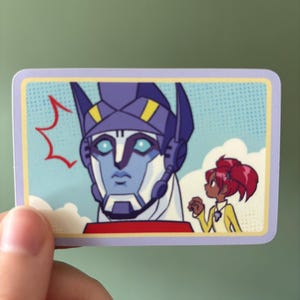 May include: A rectangular sticker featuring a cartoon illustration of a robot's face and a girl. The robot is blue and white with yellow accents. The girl has red hair and is wearing a yellow shirt. The background is light blue with white clouds.