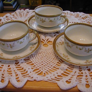 May include: Three white porcelain teacups with gold trim and floral patterns on a white crocheted doily.