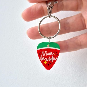 Può includere: Un portachiavi in acrilico rosso, verde e bianco a forma di fragola. Il portachiavi ha un anello e un moschettone argentati. Le parole "Viva la vida" sono scritte in bianco sulla fragola.