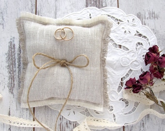 May include: A square beige linen ring pillow with frayed edges, featuring a central jute twine bow tied around the cushion. Two plain gold bands rest on the top surface of the pillow. The cushion is accented with a white lace doily and dried roses.