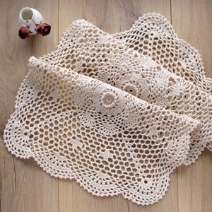 Pode incluir: Um tapete de crochê de cor creme com uma borda festonada. O tapete está dobrado ao meio e colocado sobre uma superfície de madeira.