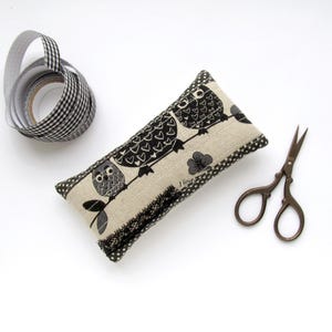 May include: A rectangular beige pin cushion with a black and white owl design, black polka dot trim, and black lace. A roll of black and white checkered ribbon and small brown scissors are also present.