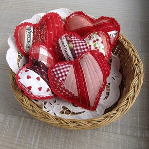 Könnte beinhalten: Eine Sammlung handgefertigter Stoffherz-Ornamente in einem Weidenkorb. Die Herzen weisen verschiedene rote und rosa Muster auf, darunter Punkte, Vichy-Karos und Blumendrucke. Einige Herzen haben Textetiketten. Der Korb steht auf einer weißen Spitzendecke.