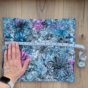 May include: A rectangular fabric with a floral pattern in shades of blue, teal, black, and pink. A measuring tape shows the fabric is approximately 16 inches wide. The fabric is on a wooden surface.