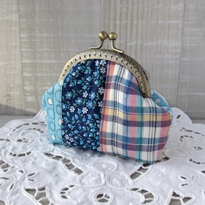 Puede incluir: Una cartera de monedas de tela de patchwork azul y blanco con un cierre de latón. La cartera está sobre un encaje blanco.