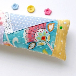 Może przedstawiać: Poduszka na szpilki w patchworku z kwiatowym wzorem w odcieniach różowego, żółtego i niebieskiego. Poduszka jest wypełniona wypełnieniem i ma żółtą obwódkę z tkaniny. Jeden szpilka jest wbita w poduszkę.