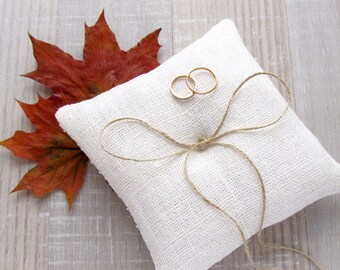 May include: A white linen ring pillow with a brown twine bow and two gold wedding bands on top. A red maple leaf is in the background.
