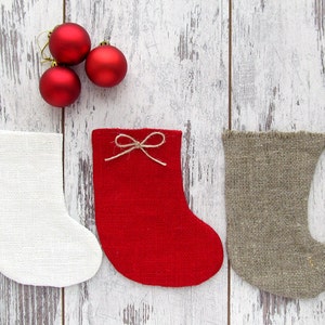 May include: Three Christmas stockings in white, red, and brown fabric. The stockings are hanging on a white wooden background. There are three red Christmas ornaments on the background.