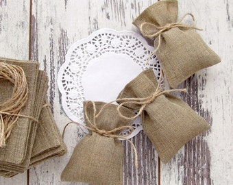 Könnte beinhalten: Eine Sammlung von Jutegeschenktüten, kleinen Jutequadraten und Bindfäden auf einer verwitterten weißen Holzoberfläche. Die Beutel sind mit Bindfaden-Schleifen gebunden. Eine weiße Papier-Spitze ist ebenfalls vorhanden.