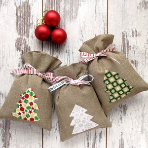 May include: Three burlap gift bags with Christmas tree appliqu&eacute;s and red and white striped ribbon ties. The trees feature various patterns and colors. Three red Christmas ornaments are in the background, all on a white wooden surface.