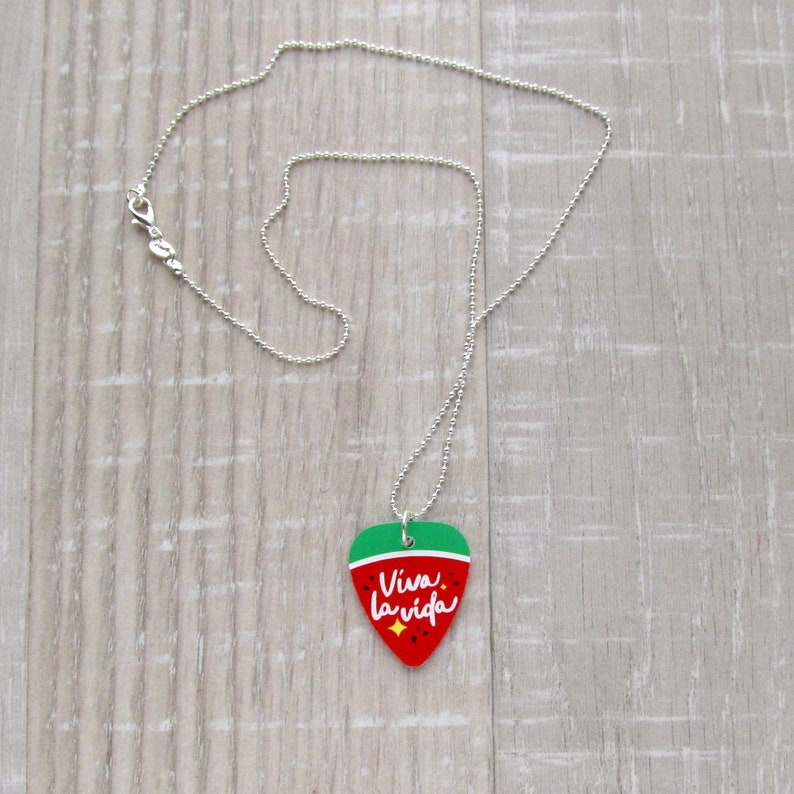 May include: A silver-colored ball chain necklace with a guitar pick pendant. The pendant is red with a green top and features the words "Viva la vida" in white script. The pendant is shaped like a heart.