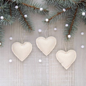 Puede incluir: Tres adornos de coraz&oacute;n de tela blanca colgando de una rama de abeto azul sobre un fondo de madera blanca.