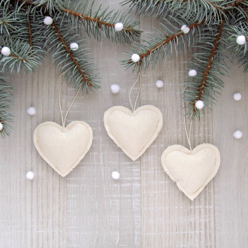 Puede incluir: Tres adornos de coraz&oacute;n de tela blanca colgando de una rama de abeto azul sobre un fondo de madera blanca.