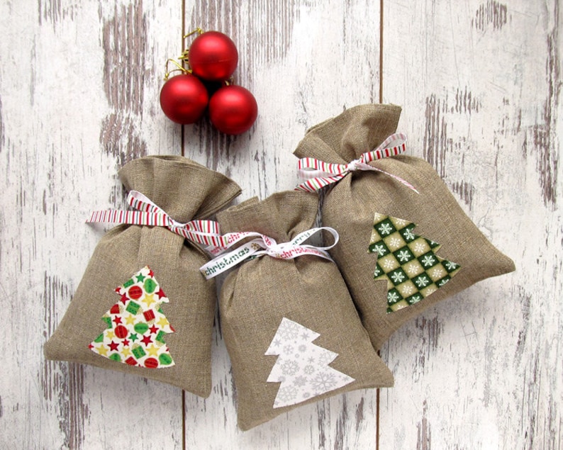 May include: Three burlap gift bags with Christmas tree appliqu&eacute;s and red and white striped ribbon ties. The trees feature various patterns and colors. Three red Christmas ornaments are in the background, all on a white wooden surface.