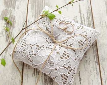 May include: White lace ring pillow with a brown twine bow and two gold wedding bands on top. The pillow is on a white wooden background with a green branch.