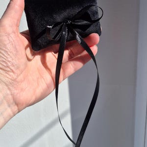 May include: A small, rectangular black velvet pillow featuring a soft, plush texture. The pillow is adorned with a thin, shiny black satin ribbon tied into a decorative bow at the center, with two long ribbon ends hanging down from the knot.