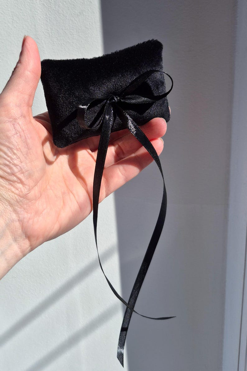 May include: A small, rectangular black velvet pillow featuring a soft, plush texture. The pillow is adorned with a thin, shiny black satin ribbon tied into a decorative bow at the center, with two long ribbon ends hanging down from the knot.