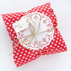 May include: A small square package wrapped in red paper with white polka dots. The package is tied with brown twine and topped with a white paper doily and a small rectangular white gift tag with the handwritten text thank you! in black ink.