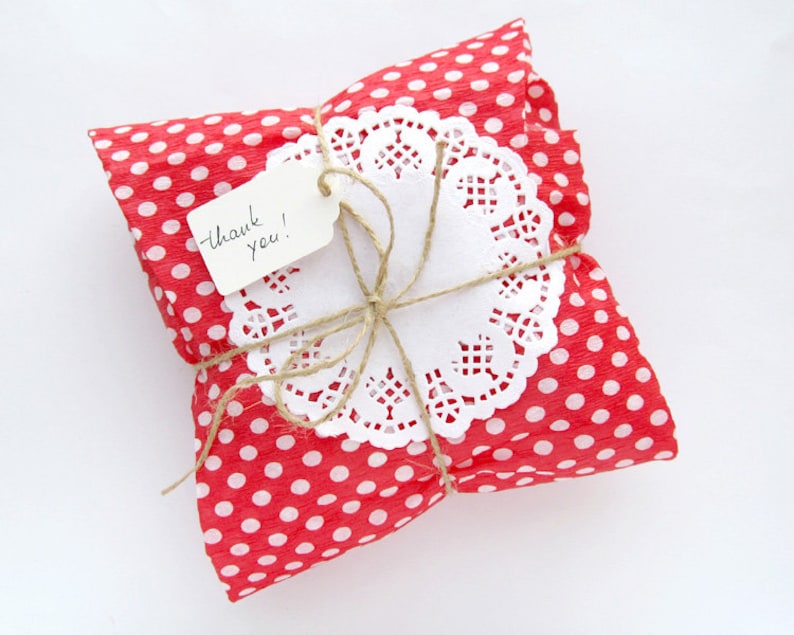 May include: A small square package wrapped in red paper with white polka dots. The package is tied with brown twine and topped with a white paper doily and a small rectangular white gift tag with the handwritten text thank you! in black ink.