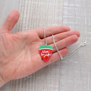 May include: A silver chain necklace with a pendant shaped like a watermelon slice. The pendant is red with green and white accents and the words "Viva la vida" in white script.