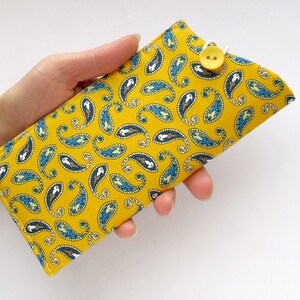 May include: A rectangular fabric case featuring a vibrant mustard yellow background with a repeating blue, white, and brown paisley pattern. The case is secured with a white elastic loop that fastens over a single yellow button at the top edge.