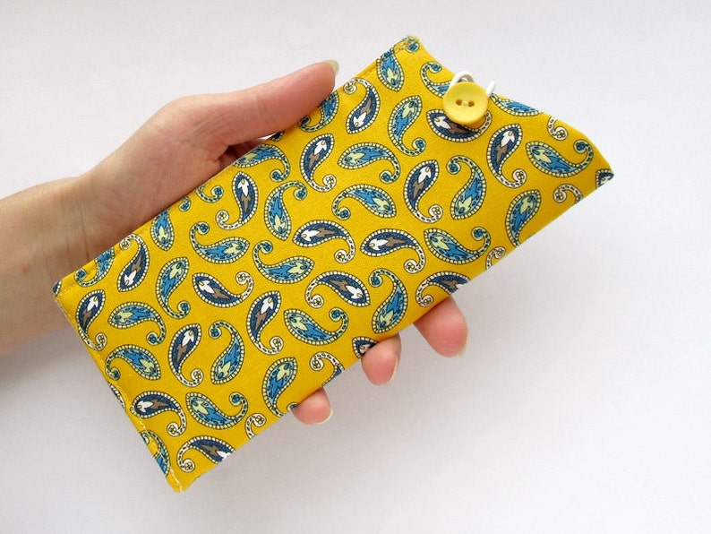 May include: A rectangular fabric case featuring a vibrant mustard yellow background with a repeating blue, white, and brown paisley pattern. The case is secured with a white elastic loop that fastens over a single yellow button at the top edge.
