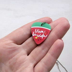 May include: A guitar pick-shaped pendant with a green top and red bottom, featuring the words "Viva la vida" in white script. The pendant has a silver loop and is attached to a silver chain. The design includes yellow star accents.